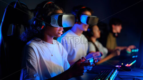 Black teen smiling girl in VR headset playing video games using gamepad in video game club with blue illumination - Starpik