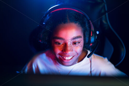 Black teen smiling girl in headset playing video games in video game club with blue and red illumination - Starpik