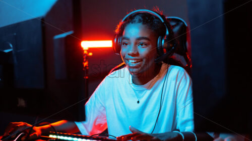 Black teen smiling girl in headset playing video games in video game club with blue and red illumination. Keyboard with illumination - Starpik