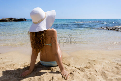 Blonde woman in turquoise swimsuit with white hat sitting on sand and looking to blue sea - Starpik