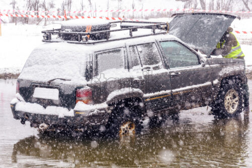 Broken jeep in middle of a river with smoke coming from motor. Driver repairing. Rally competition with mud and snow. Spectators looking from hill. Magdacesti, Moldova - Starpik