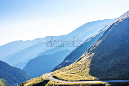 Daylight aerial view from top to winding road with car riding on it. Silhouette of Mountains in fog on background. Bright blue clear sky. Negative copy space, place for text. Transfagarasan, Romania - Starpik