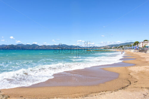 Daylight sunny view to beachline and sea of resort. People relaxing on sand. Buildings and big mountains on background. Negative copy space. Bright blue sky with clouds, Cannes, France - Starpik