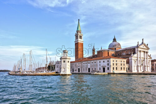 Daylight view from boat to San Giorgio Maggiore church with ornamented facade. Little port with parked boats and bright blue sky with clouds on background. Place for text. Venice, Italy - Starpik