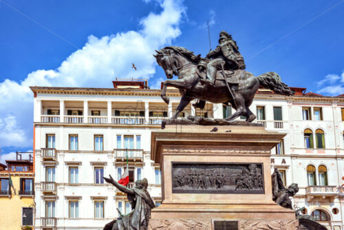 Daylight view from bottom to Equestrian Statue of King Victor Emmanuel II in Riva degli Schiavoni waterfront. Historic architecture hotel on background. Bright blue sky with clouds Venice, Italy - Starpik