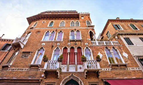 Daylight view from bottom to old brick ornamented facade of a building with balconies. Bright blue cloudy sky on background. Venice, italy - Starpik