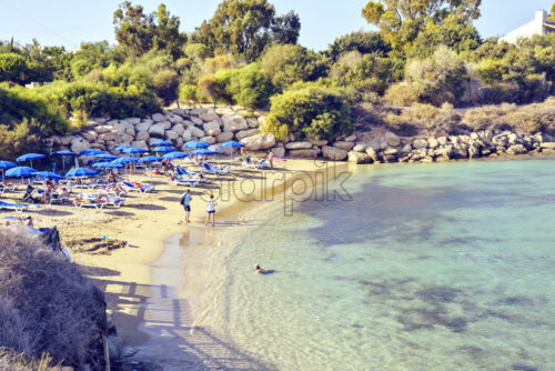 Daylight view to beach with people relaxing in water and sunbathing on seashore. Green trees and rocks on background. Negative copy space, place for text. Protaras, Cyprus - Starpik