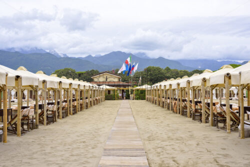 Daylight view to beige sun chairs and sunshades. People walking on background. Cloudy sky and mountains on background. Negative copy space, place for text. Forte dei Marmi, Italy - Starpik