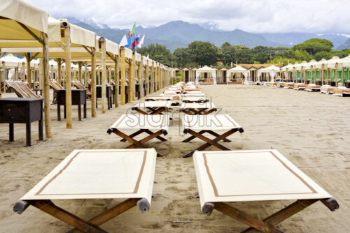 Daylight view to beige sunchairs and sunshades on beach. Changing rooms, big mountains and cloudy sky on background. Negative copy space, Place for text, Forte dei marmi, Italy - Starpik