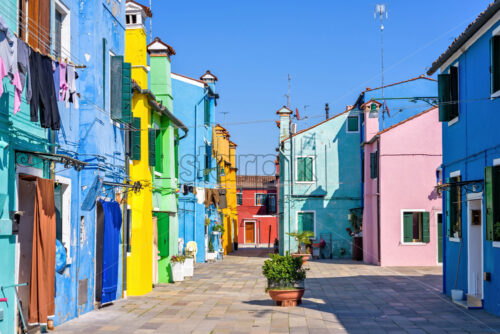 Daylight view to colorful vibrant housefront facades with clothes drying outside. Shadows and sun shining. Negative copy space, place for text. Bright blue clear sky. Burano, Italy - Starpik