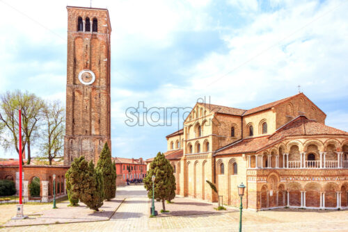 Daylight view to historic architecture building of Basilica Santa Maria e San Donato with clock watch tower on Murano Island. Bright blue sky with clouds. Warm mood. Venice, Italy - Starpik