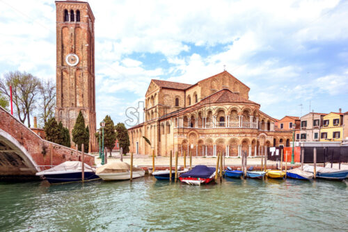 Daylight view to parked boats and historic architecture building of Basilica Santa Maria e San Donato with clock watch tower on Murano Island. Bright blue sky with clouds. Warm mood. Venice, Italy - Starpik