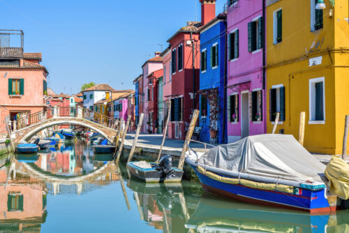 Daylight view to parked boats in a canal with image of bridge reflecting on water. Colorful buildings and bright blue clear sky. Negative copy space, place for text. Burano, Italy - Starpik