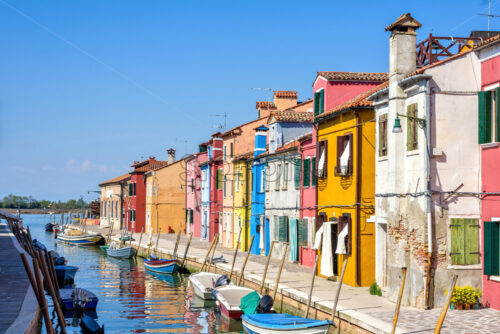 Daylight view to parked boats in a canal with image reflecting on water. Colorful buildings and bright blue clear sky. Negative copy space, place for text. Burano, Italy - Starpik