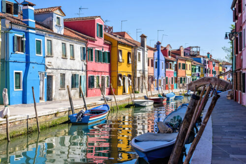 Daylight view to parked boats in a canal with image reflecting on water. Colorful buildings and bright blue clear sky. Negative copy space, place for text. Burano, Italy - Starpik