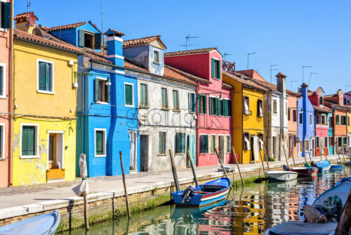 Daylight view to parked boats in a canal with image reflecting on water. Colorful buildings and bright blue clear sky. Negative copy space, place for text. Burano, Italy - Starpik