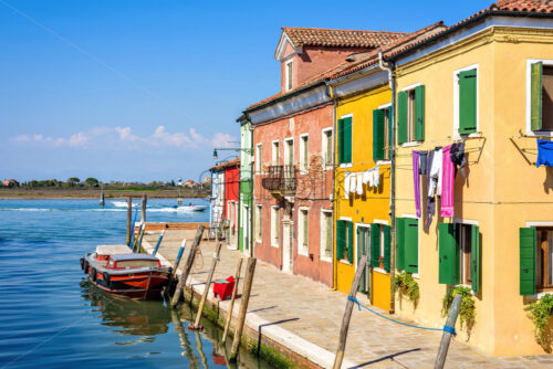 Daylight view to parked boats in a canal with image reflecting on water. Colorful buildings and bright blue clear sky. Negative copy space, place for text. Burano, Italy - Starpik