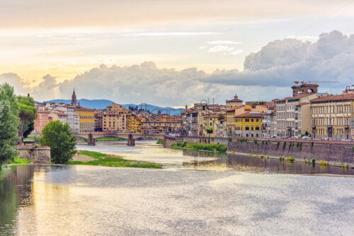 Daylight view to sunset above Arno river with reflections. Green trees, city buildings, bridge and Cathedral Saint Mary of the Flower on background. Florence, Italy - Starpik