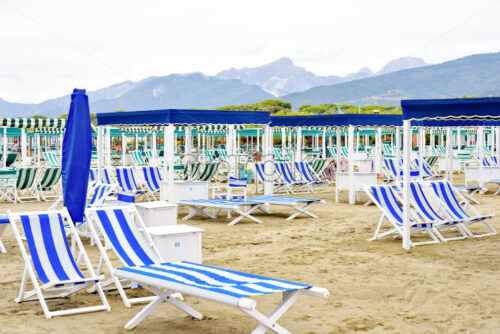 Daylight view to vibrant blue sunchairs and sunshades on beach. Cloudy sky, buildings and mountains on background. Negative copy space, place for text. Forte dei Marmi, Italy - Starpik