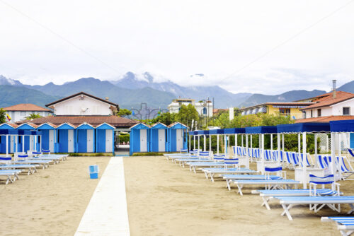 Daylight view to vibrant blue sunchairs and sunshades on beach. Cloudy sky, buildings and mountains on background. Negative copy space, place for text. Forte dei Marmi, Italy - Starpik