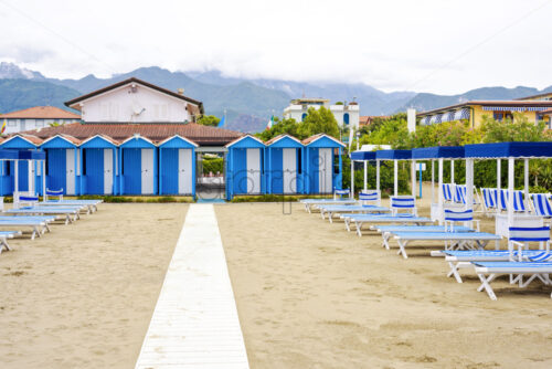 Daylight view to vibrant blue sunchairs, changing rooms and sunshades on beach. Cloudy sky, buildings and mountains on background. Negative copy space, place for text. Forte dei Marmi, Italy - Starpik