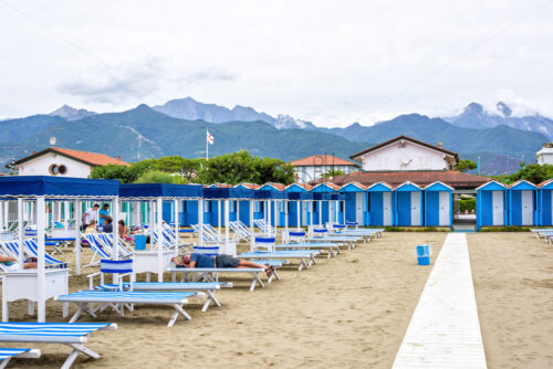 FORTE DEI MARMI, ITALY – JUNE 30, 2017: Daylight view to beach with people resting on sunchairs in a cloudy day. Big mountains and sky on background. Negative copy space, place for text. - Starpik