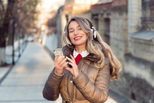 Happy woman listening to music on wireless headphones connected to mobile phone, on the streets at sunset - Starpik