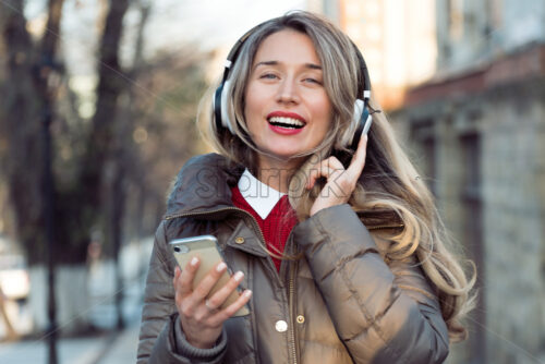 Happy woman listening to music on wireless headphones connected to mobile phone, on the streets at sunset - Starpik