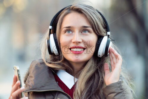 Happy woman listening to music on wireless headphones connected to mobile phone, on the streets at sunset - Starpik