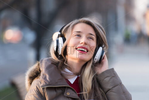 Happy woman listening to music on wireless headphones connected to mobile phone, on the streets at sunset - Starpik