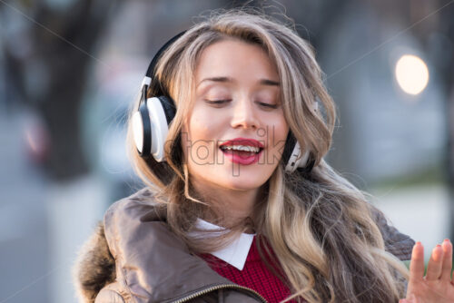 Happy woman listening to music on wireless headphones connected to mobile phone, on the streets at sunset - Starpik