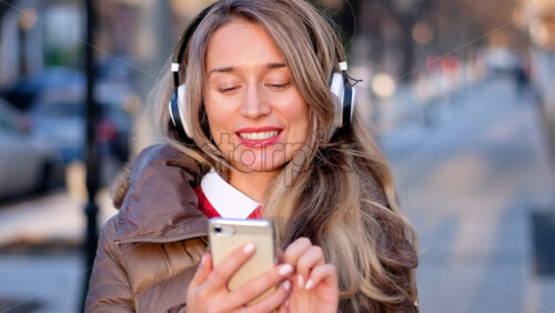 Happy woman listening to music on wireless headphones connected to mobile phone, on the streets at sunset - Starpik