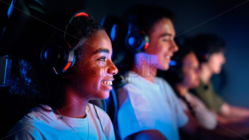 Multiracial group of teens in headsets playing video games in video game club with blue and red illumination. Keyboards and mice with illumination - Starpik