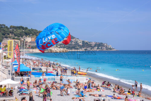 NICE COTE D’AZUR, FRANCE – JUNE 27, 2017: Beautiful daylight view to beach of Nice Cote d’Azur. People relaxing on beach and swimming in water. Parachute man landed on sand. - Starpik