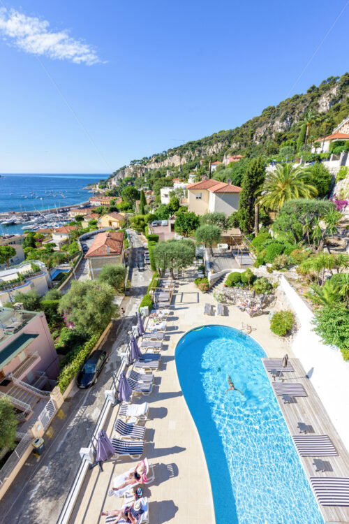 NICE, FRANCE – JUNE 28, 2017: Daylight view to hotel pool with people sitting on armchairs. Woman swimming in water. Yachts cruising on water - Starpik