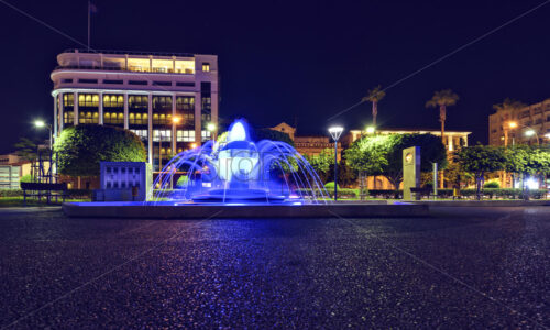 Night view to modern public fountain with blue light glowing. Hotel and park on background. Negative copy space, place for text. Molos Promenade, Limassol, Cyprus - Starpik