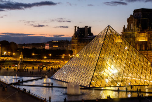 PARIS, FRANCE – OCTOBER 03, 2018: Night shot of Louvre museum in Paris. Orange sky in the background. Reflection of a pyramid on the water - Starpik