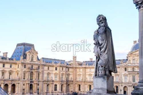 PARIS, FRANCE – OCTOBER 03, 2018: Statue representing an old man in the Louvre museum’s teritory. Magenta accents on the building. Clear sky at sunset - Starpik