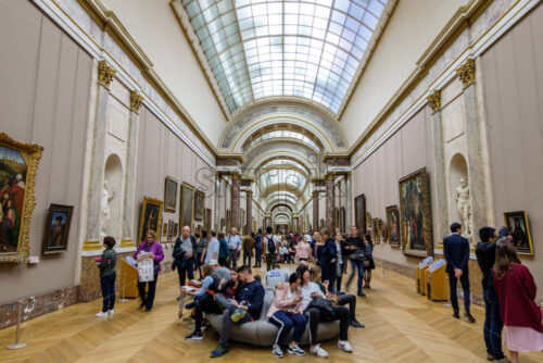 PARIS, FRANCE – OCTOBER 03, 2018: Tourists in Louvre museum having a rest on the couches. Long tunnel in the background full of people. Artworks on the walls - Starpik