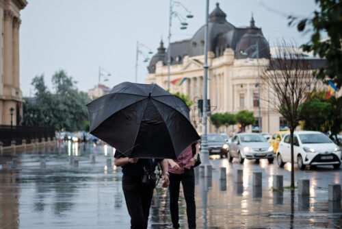 People with black umbrella in the rain at Calea victoriei square with buildings in bucharest, romania - Starpik