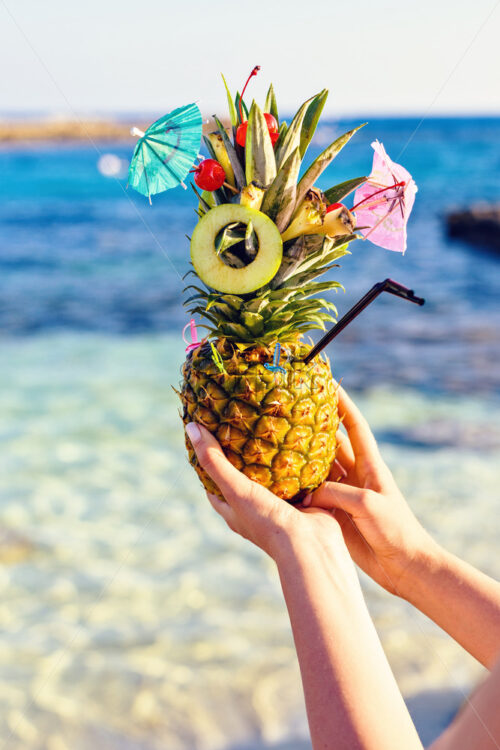 Pineapple decorated in a great exotic style with fruits and a mini umbrella. The fruit is being hold by two woman hands. Sea on background. Nissi Beach, Ayia Napa, Cyprus. - Starpik