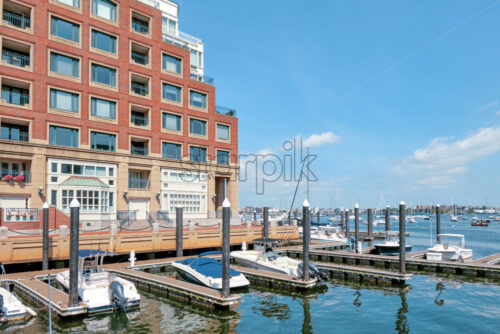 Shot of several moored and floating boats on the smooth surface of the water in Boston with beautiful residential building and blue clear sky in the background - Starpik