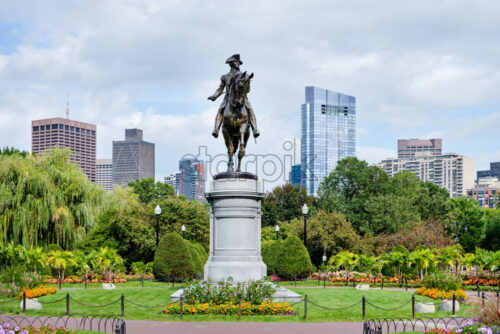 Shot of the George Washington Monument located in Boston Public Garden full of greenery with a lot of high modern buildings in the background. The weather is cloudy - Starpik