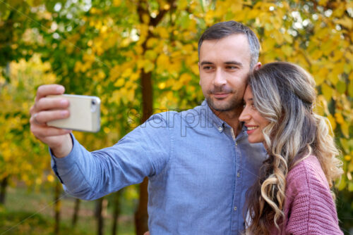 Shot of the beautiful smiling couple taking selfie with autumn yellow and green leaves in the background - Starpik