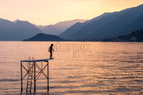 Silhouette of a woman trying to jump into water from platform. Lake Como with mountains at sunset. Cloudy sky. View from Bellagio, Italy - Starpik