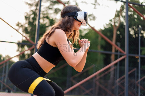 Smiling woman in VR headset in a tracksuit doing exercises with elastic band on a sports field in a park - Starpik