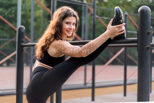 Smiling woman in a tracksuit doing exercises on a sports field in a park, looking into the camera - Starpik