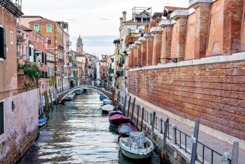 Sunset view to canal with parked boat, bridges and colorful buildings. Purple sky with clouds reflecting on water. Venice, Italy - Starpik