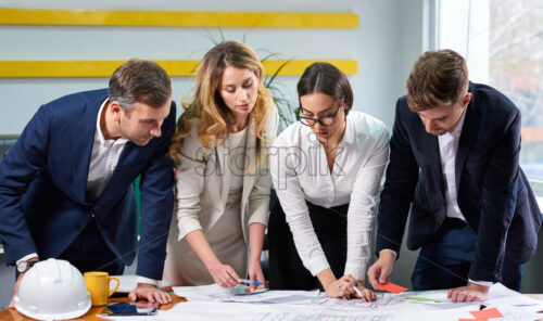 Team of mature women and men at meeting table discussing a construction plan blueprint with hard hat in office - Starpik