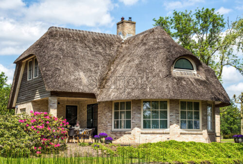 Traditional house with rustic thatched roof in a sunny day. Trees, flowers and bright blue sky with clouds on background. Giethoorn, Netherlands - Starpik